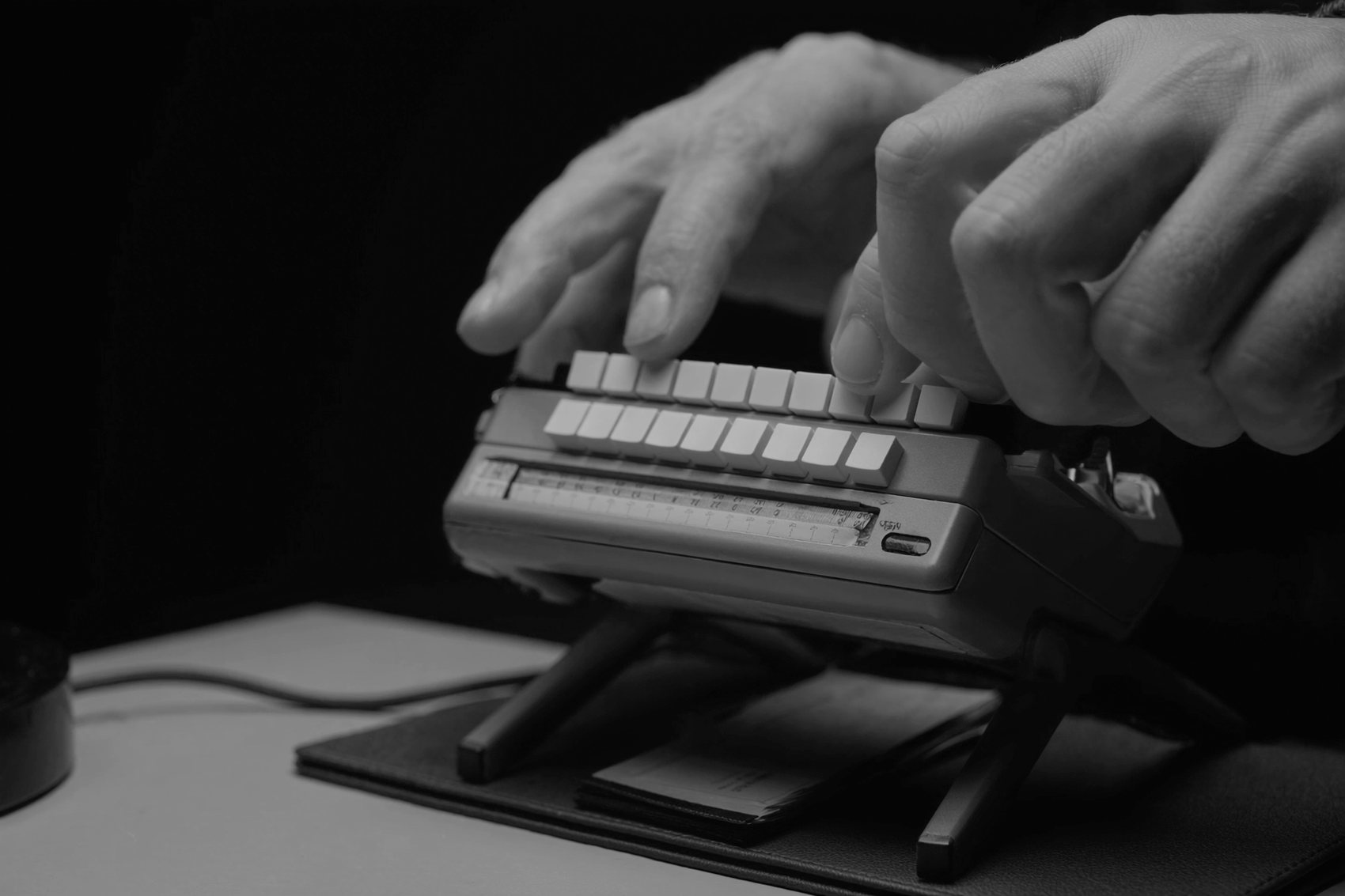 Two hands on the flat blank keytops of a stenotype machine, its fanfold paper tray folding beneath it.
