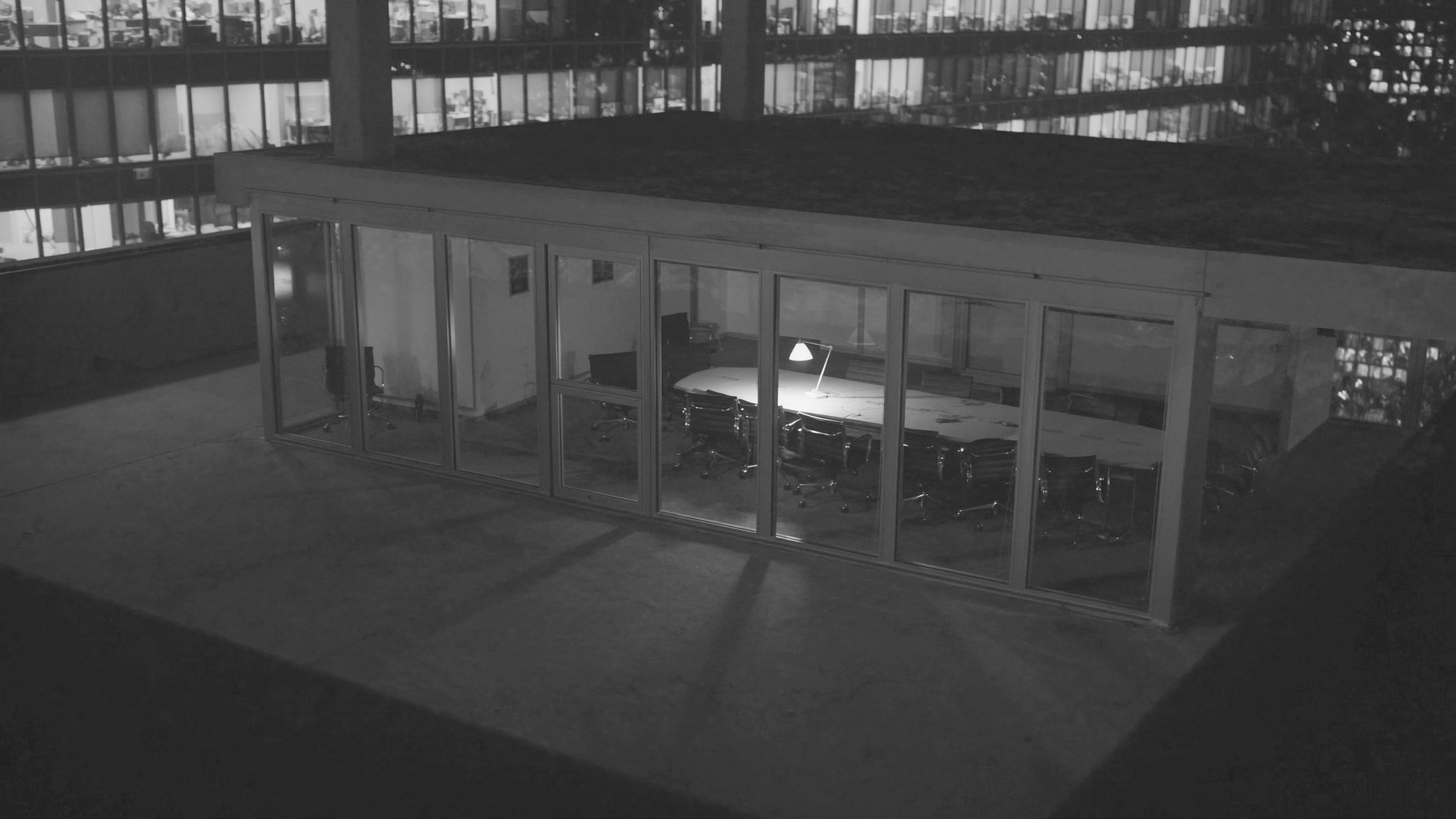 An empty glass-walled conference room at night, one lamp still burning over papers left on the table, lit windows in the tower opposite.