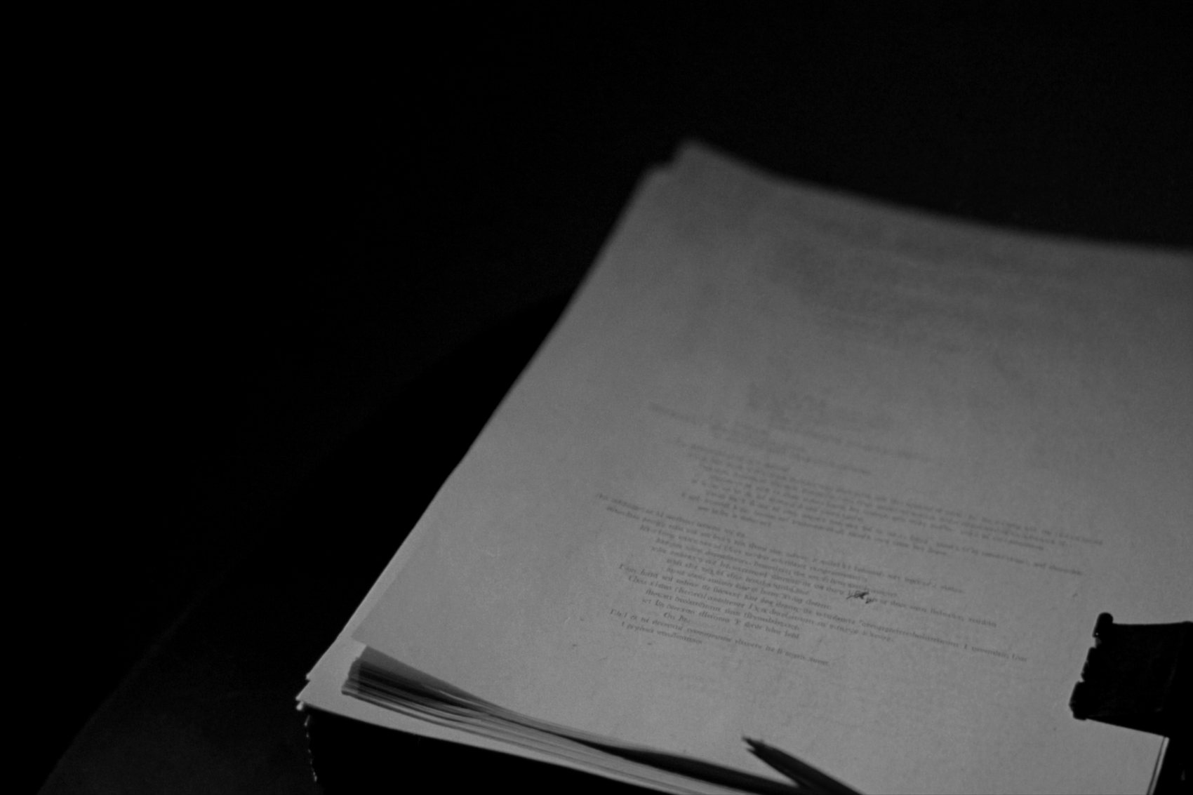 A thick stack of printed pages under a desk lamp, the top page too softly focused to read, a red pen across the corner.