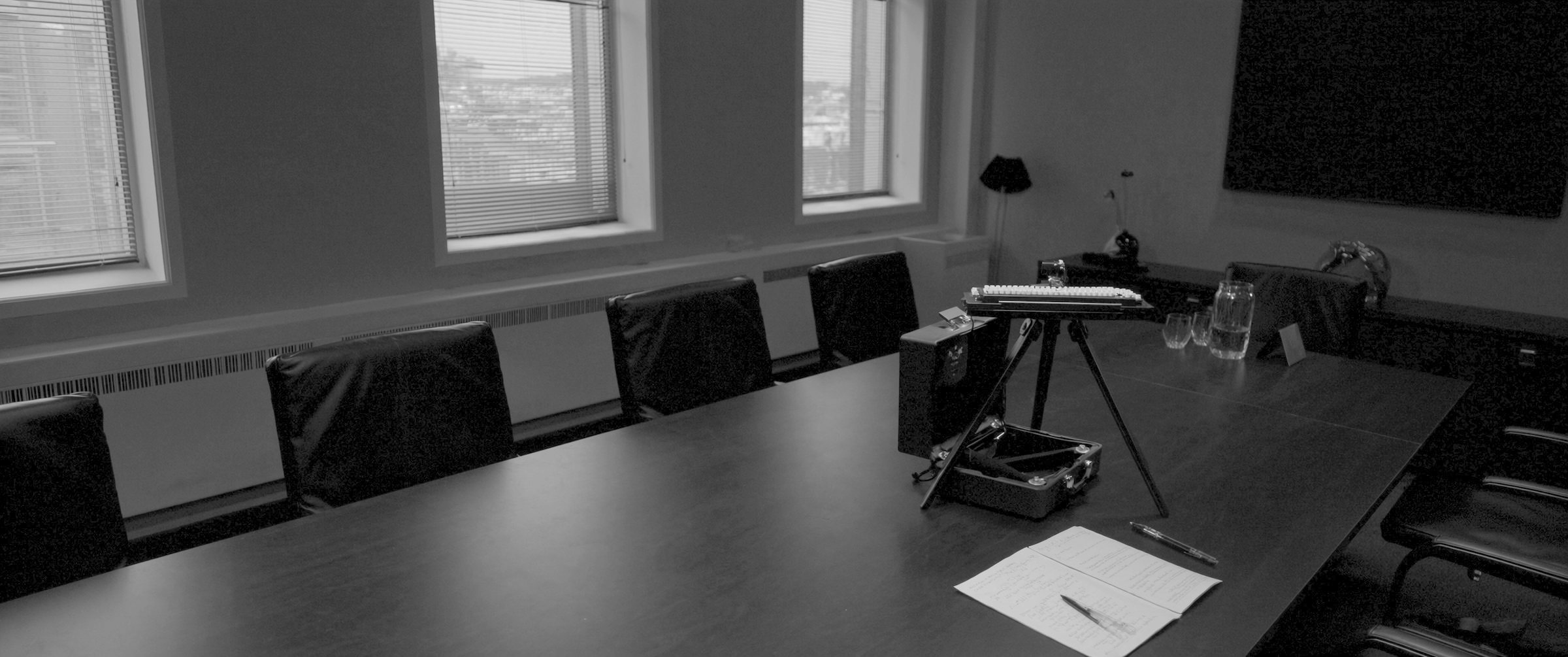 A stenotype machine on a tripod at the end of a long empty conference table, chairs pushed in, blinded windows behind it.