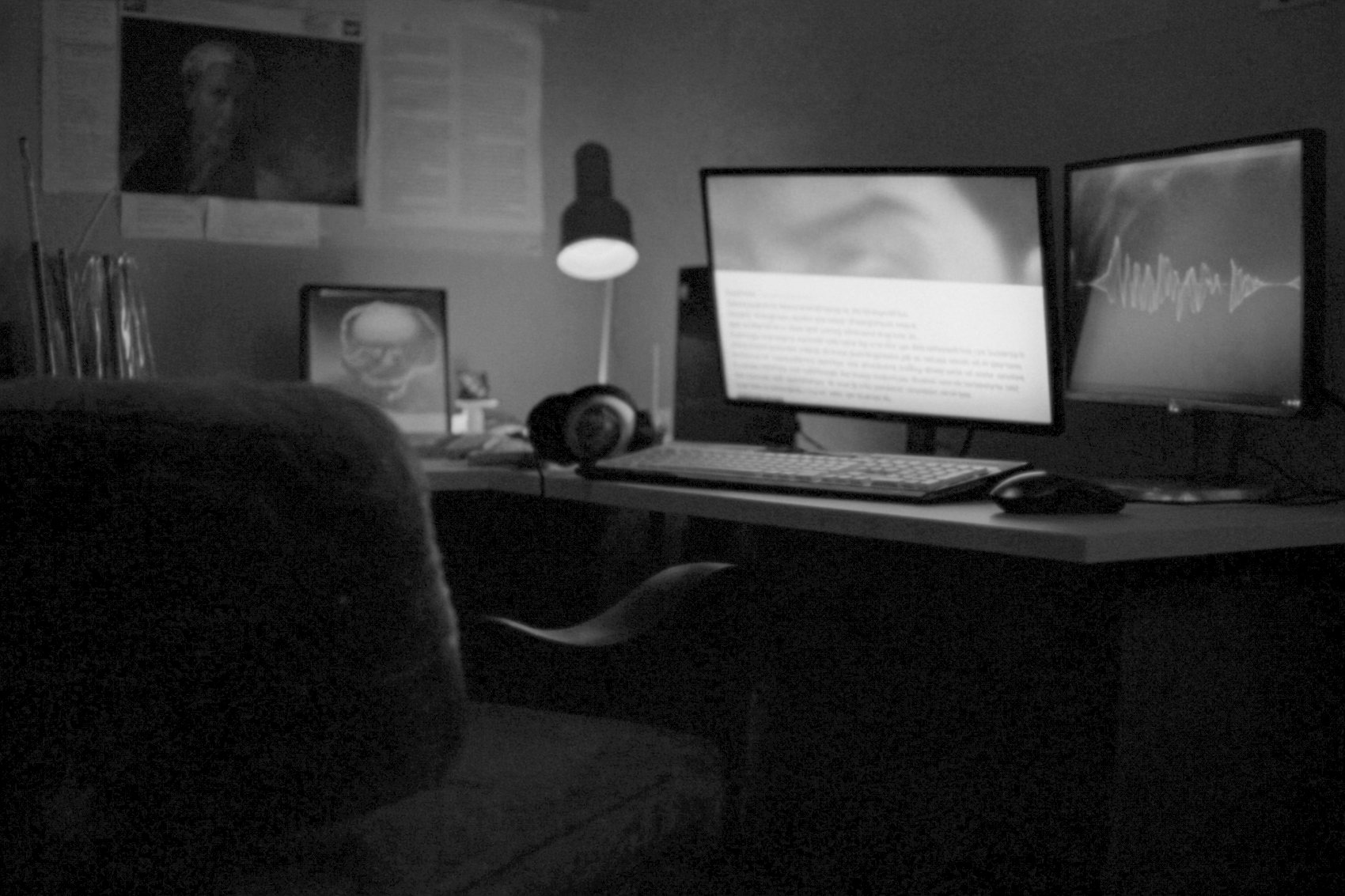 A dark room lit by two monitors, one running an audio waveform and one a page of text over a paused video frame, headphones on the desk.